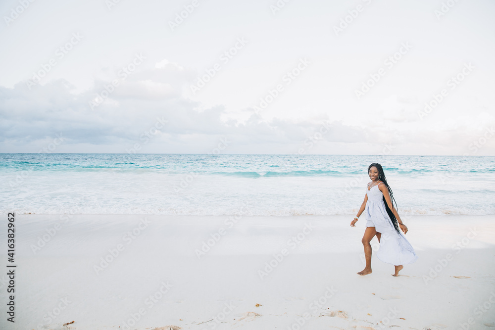 Young black woman having fun at seaside. She is twenty years old, mixed race caucasian and african black, with curly and voluminous hair, running with open arms and happy face.