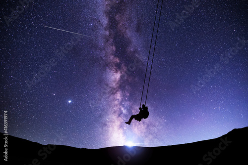 Fototapeta Naklejka Na Ścianę i Meble -  Fantasy night scene. Silhouette of a guy swinging on a swing under bright milky way galaxy. Beautiful starry night.