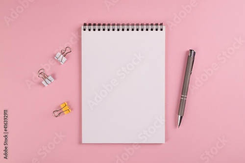 A notebook with a blank page, a ballpoint pen and multi-colored clips for paper lie on a pink background. Copy space, layout, mockup.