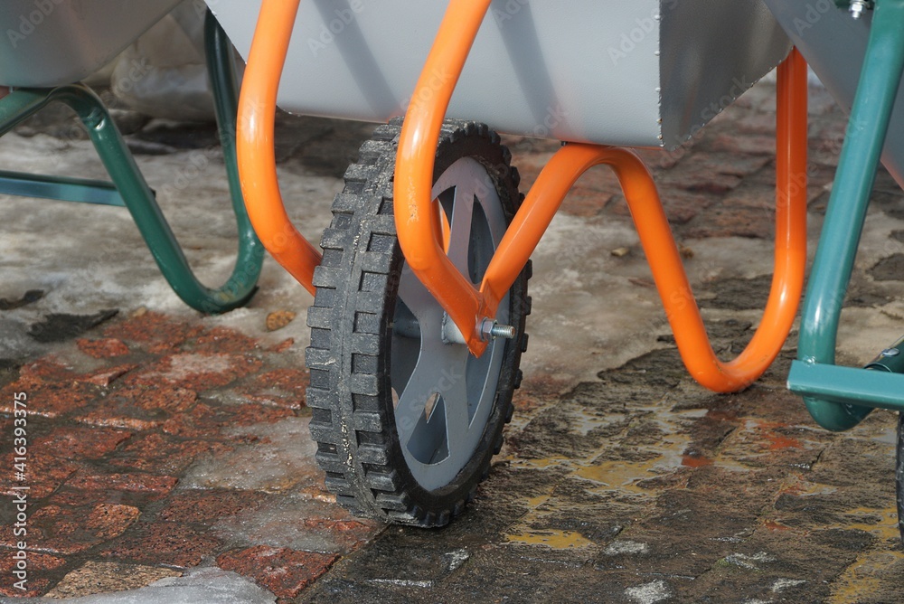  a black wheel on a orange metal garden wheelbarrow stands on a brown sidewalk and white snow on a winter street