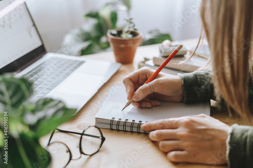 Distance learning online education and work. Business woman with plants writes in notebook. Cropped freelance girl with laptop at home office. Using computer and online shops.