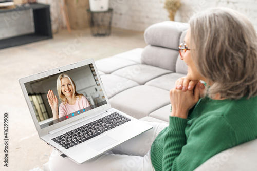 Back view on the laptop screen with a video call participant on it, two middle-aged women have a video meeting on the laptop. Senior lady talks online with an adult daughter, mid-age friend, coworker