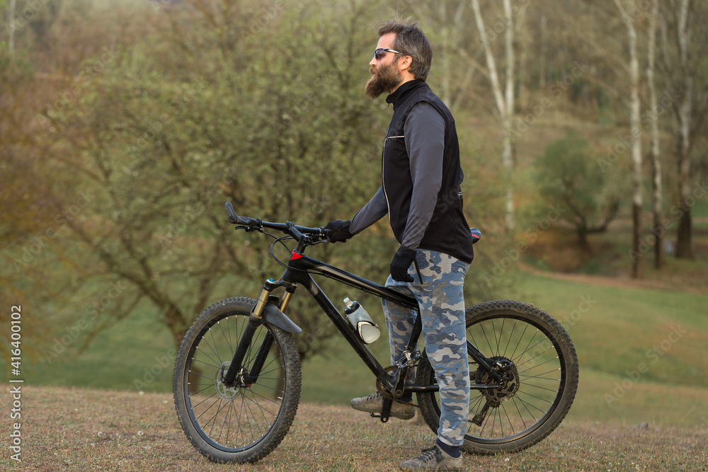 Sports brutal bearded guy on a modern mountain bike. Cyclist on the green hills in the spring.