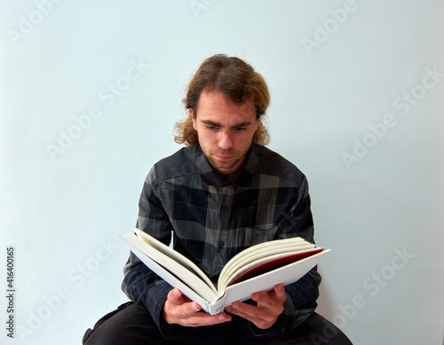 A handsome Caucasian man with long hair reading a book on the background of the white wall