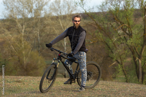 Sports brutal bearded guy on a modern mountain bike. Cyclist on the green hills in the spring.