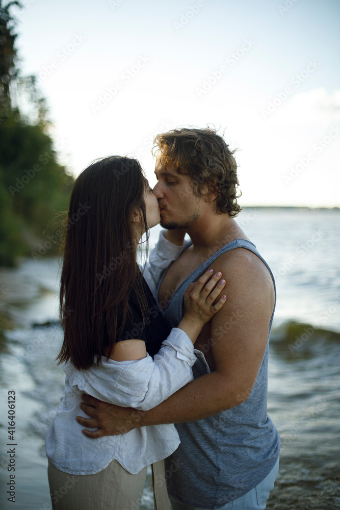 young couple a brunette girl and a curly guy kiss passionately in french on the seashore at sunset. love story on the sea beach