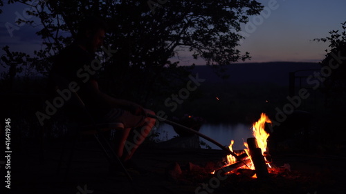 The man straightens the coals in the fire. The fire burns in the night against the background of the river and mountains.