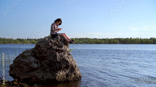 A young woman sits on a stone by the river with a phone in her hands.