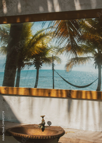 Modern luxury summer holiday or vacation beach house exterior public area close to the sea with an old stylish sink and a hammock tied to palm trees viewed through the mirror.