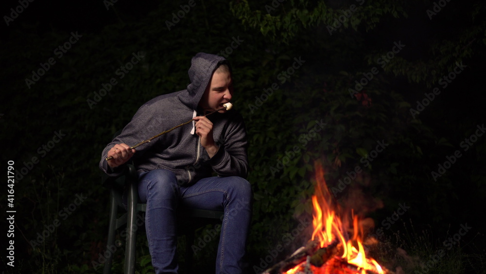 Obraz premium A young man in a hood fries marshmallows over a fire. Campfire at night.