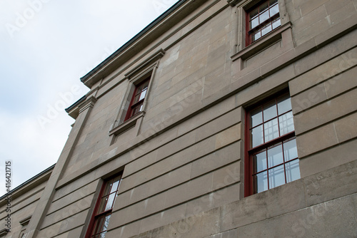 The exterior of a vintage limestone block wall with multiple windows. The tall government building has a blue sky with clouds in the background. The trim around the double hung windows is red wood. 