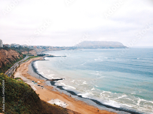 View of the beach in Lima, Peru. There are buildings on the hill of the city. The sky is cloudy and there are people walking close to the shore.
