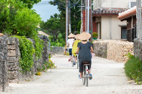 Three people riding bicycles, one after the other, strolling through the traditional village of Taketomi, wearing the traditional straw hat.