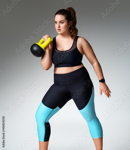 Strong woman workout with kettlebell. Photo of model with curvy figure in fashionable sportswear on grey background. Sports motivation and healthy lifestyle