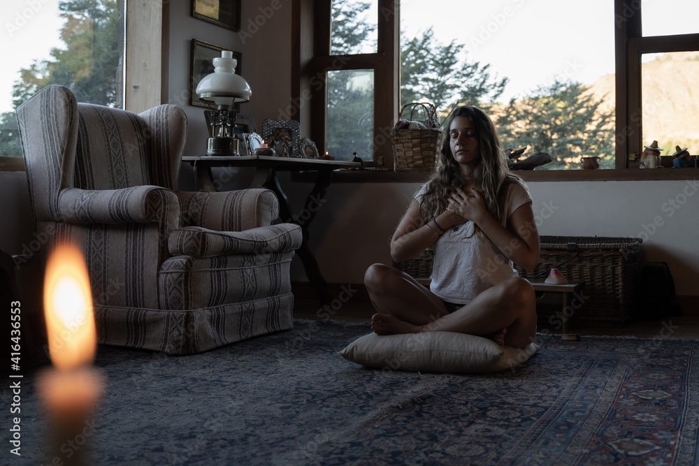 Young latin female meditating sitting on a pillow in casual indoor spiritual studio with big green window with vegetation in a wooden home