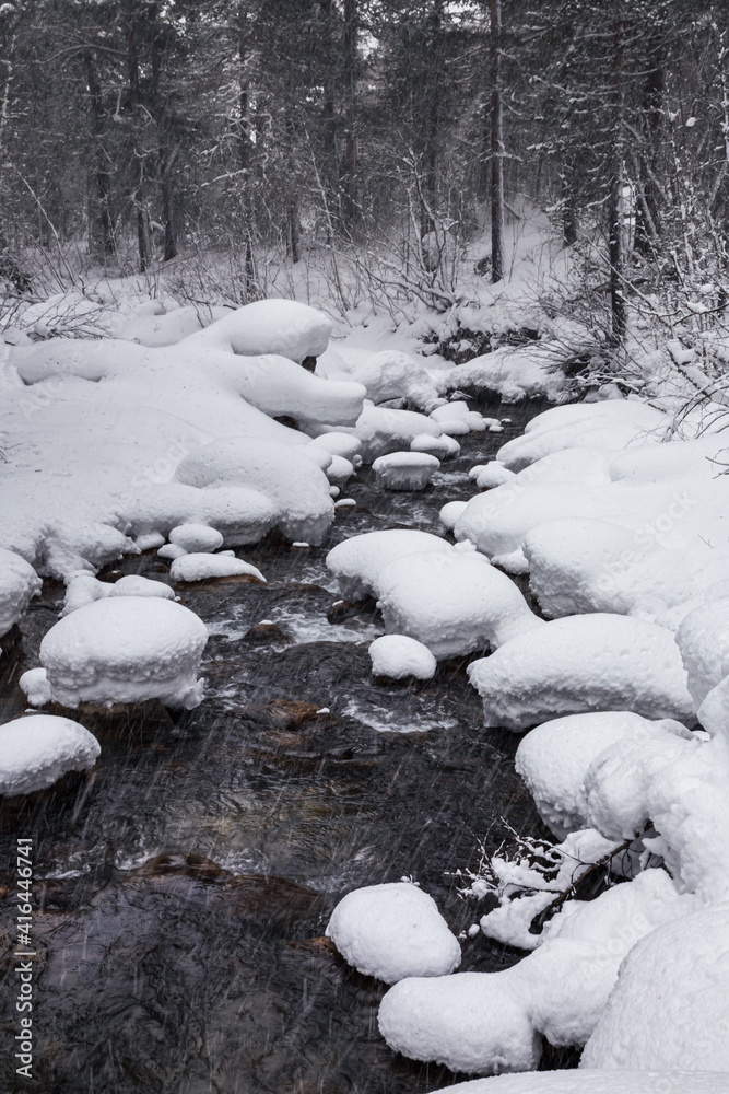 A narrow mountain river flows through a winter valley through a snow-covered forest. High caps of snow lie on stones. Heavy snowfall. Traces of flying snowflakes are visible against a dark background A narrow mountain river flows through a winter valley through a snow-covered forest. High caps of snow lie on stones. Heavy snowfall. Traces of flying snowflakes are visible against a dark background