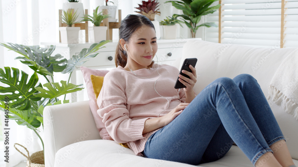 Young attractive beautiful asian woman wear earphone headphone listen to social media application clubhouse podcast or audio chat app feeling happy comfort cozy at home sitting at sofa couch.