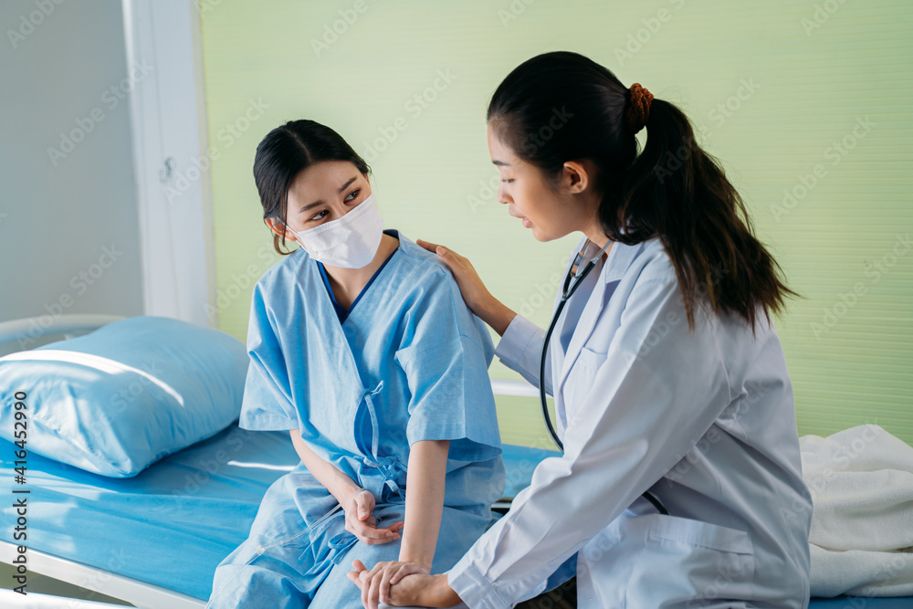 Fototapeta premium Young female doctor with stethoscope holding hand and talking to Asian patient sitting on bed in hospital wearing safety face mask