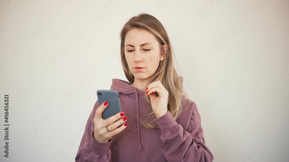 Beautiful serious blond woman in a hoodie looking at a smartphone screen over white background. Beautiful serious blond woman in a hoodie looking at a smartphone screen over white background.