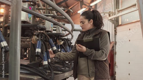Medium long of young Mixed-race female worker standing, taking notes, checking serviceability of rotating industrial milking machine 