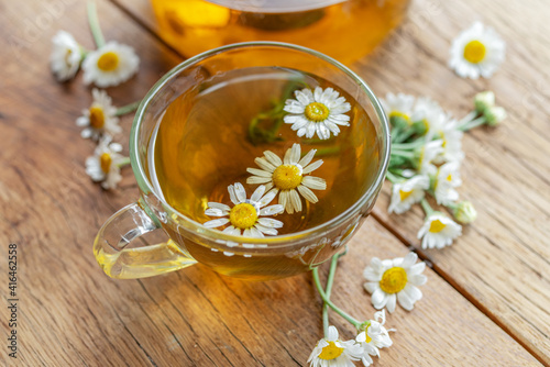 Herbal chamomile tea and chamomile flowers near teapot and tea glass on wooden table. Countryside background.