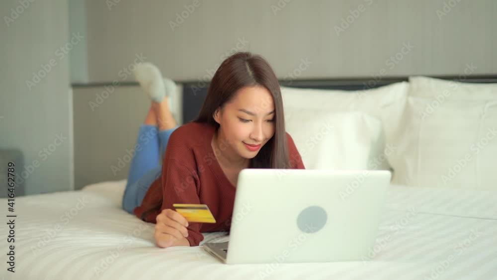 Happy Asian woman lying on bed with credit card and laptop. Slow-motion Happy Asian woman lying on bed with credit card and laptop. Slow-motion