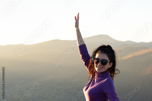 Young smiling woman raising up her hand enjoying warm sunset evening in summer mountains.