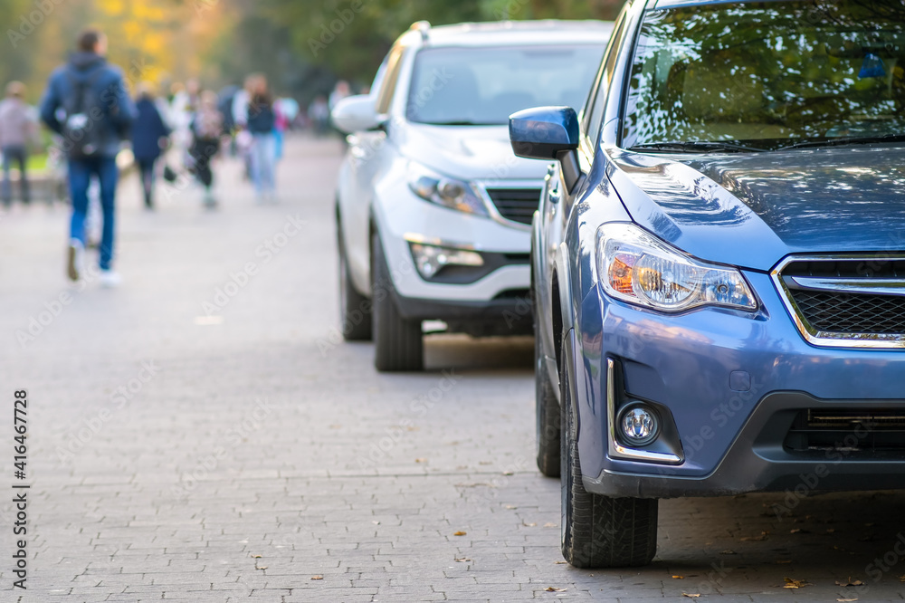 Cars parked in a row on a city street side on bright autumn day with blurred people walking on pedestrian zone. Cars parked in a row on a city street side on bright autumn day with blurred people walking on pedestrian zone.