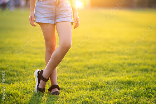 Closeup of young child girl legs in denim shorts standing on green grass lawn on warm summer evening.