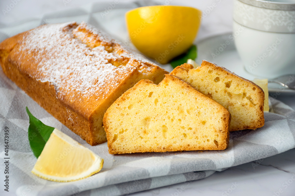 Moist lemon pound cake on kitchen towel on marble table with slices of lemon and cup of tea on plate. Delicious breakfast, traditional tea time treat. Reciepe of English lemon pie loaf. Moist lemon pound cake on kitchen towel on marble table with slices of lemon and cup of tea on plate. Delicious breakfast, traditional tea time treat. Reciepe of English lemon pie loaf.