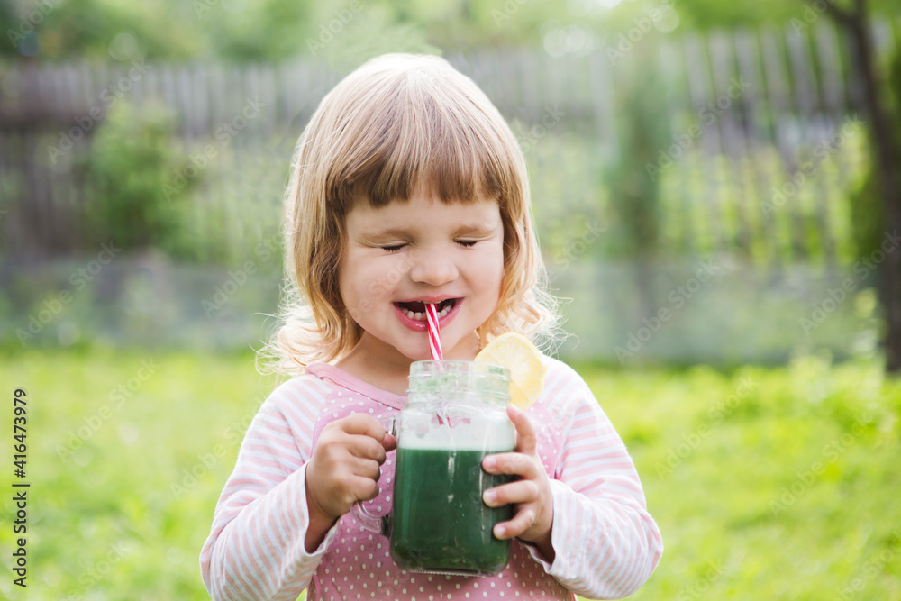 Cute little child drinks healthy green smoothie with straw in a jar mug against the background of greenery outdoor. Cute little child drinks healthy green smoothie with straw in a jar mug against the background of greenery outdoor.