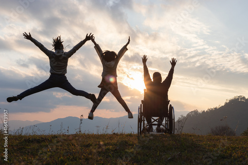 Silhouette Disabled handicapped woman in wheelchair with raised arms and children jumping at sunset. happy family concepts.