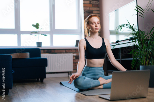 Relaxed redhead young woman with closed eyes meditating at home sitting in lotus position on yoga mat near laptop computer. Peaceful red-haired lady holding hands on knee in Om position.