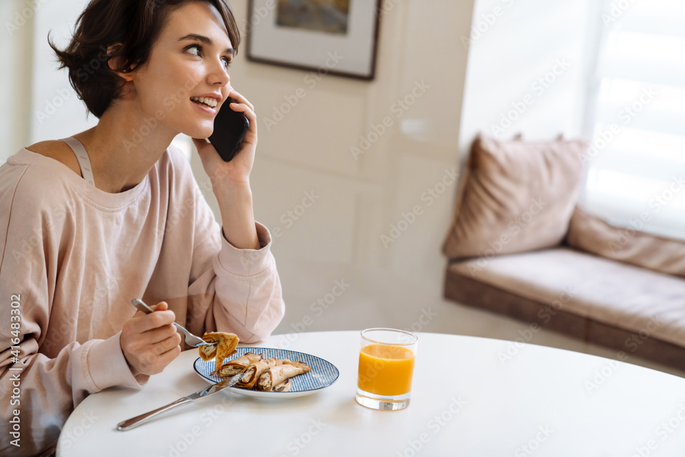 Joyful beautiful girl talking on cellphone while having breakfast