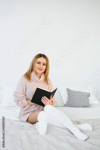 A woman reading a book and smiling as she sits in bed. The alarm clock on the desk beside her.