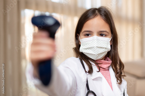 Little girl with doctor's uniform and medical mask using an infrared thermometer. Covid-19 virus safety concept.