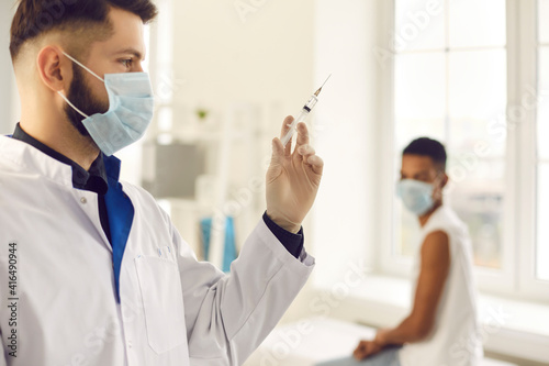 Closeup side view young male doctor in medical face mask preparing syringe for administering flu  AIDS or COVID-19 vaccine to patient against blurred hospital background with man waiting for injection