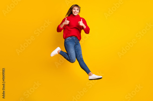 Full size profile photo of blond optimistic lady jump thumb up wear red sweater jeans sneakers isolated on bright yellow color background