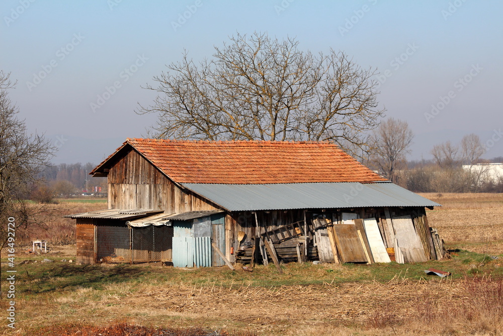 Wooden boards old barn with attached storage shed and improvised dog kennel surrounded with dry grass and trees without leaves on warm sunny winter day Wooden boards old barn with attached storage shed and improvised dog kennel surrounded with dry grass and trees without leaves on warm sunny winter day