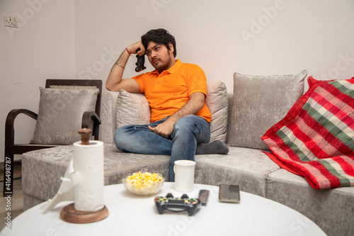 Asian Indian man gaming, streaming and watching TV alone in living room on a grey sofa and white table