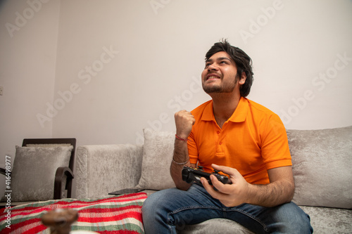 Asian Indian man gaming, streaming and watching TV alone in living room on a grey sofa and white table