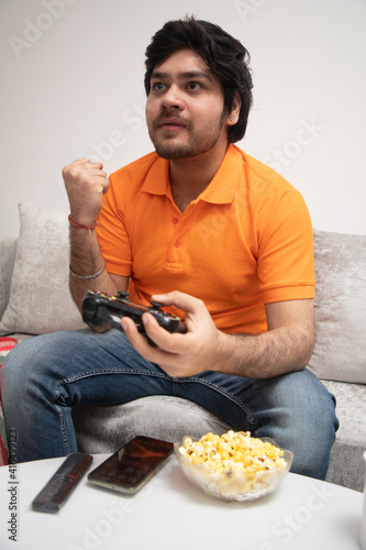 Asian Indian man gaming, streaming and watching TV alone in living room on a grey sofa and white table