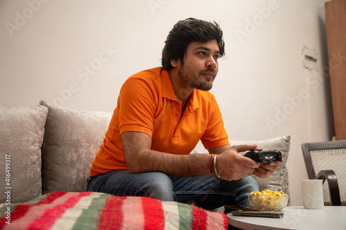 Asian Indian man gaming, streaming and watching TV alone in living room on a grey sofa and white table