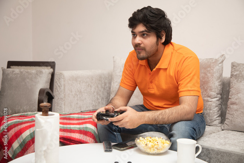 Asian Indian man gaming, streaming and watching TV alone in living room on a grey sofa and white table