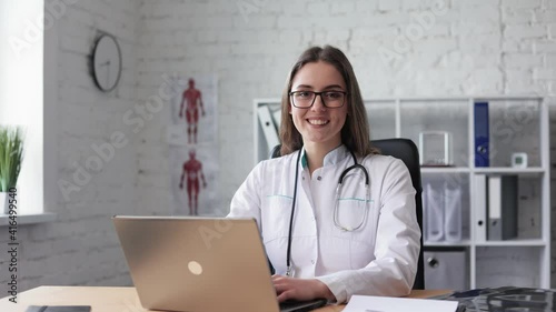 Caucasian female doctor in lab coat, glasses and stethoscope over her neck using laptop at desk, looking at camera and smiling while working in medical office. Young medical assistant. slow motion.