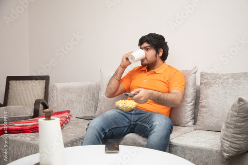 Asian Indian man gaming, streaming and watching TV alone in living room on a grey sofa and white table
