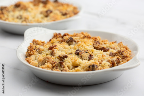 Two plates of delicious apple crumble pie isolated on marble background. Close-up of double crumble pies with fruits, nuts and cinnamon in white bowls. Dessert menu concept.