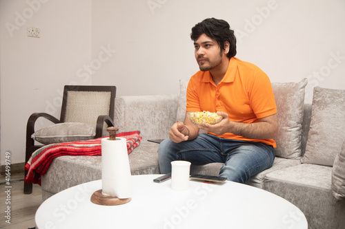 Asian Indian man gaming, streaming and watching TV alone in living room on a grey sofa and white table