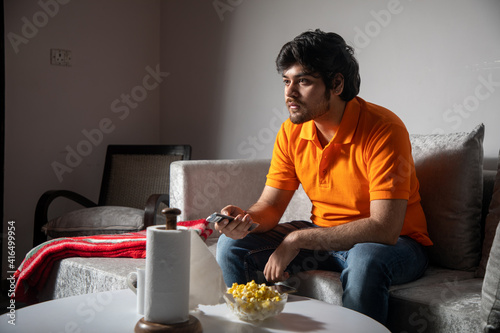 Asian Indian man gaming, streaming and watching TV alone in living room on a grey sofa and white table