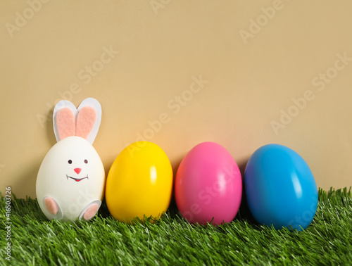 Bright eggs and white one as Easter bunny on green grass against beige background